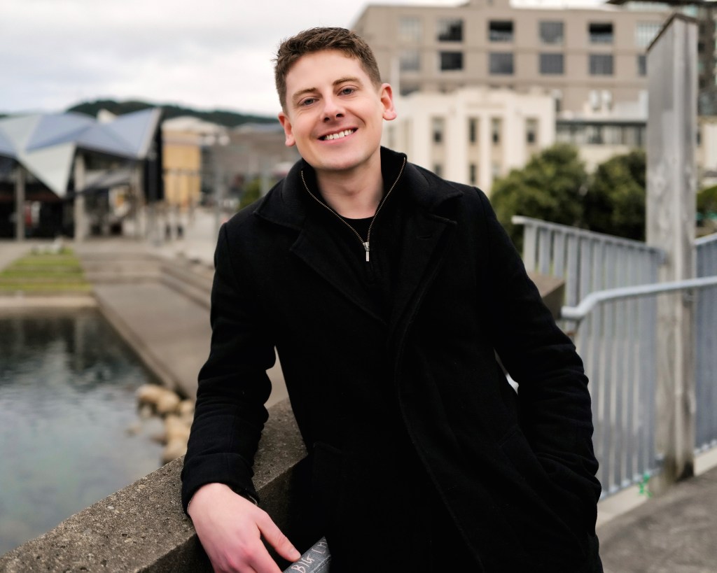 A smiling young person in a black coat leans against a stone barrier near a body of water, with modern buildings and greenery in the background.
